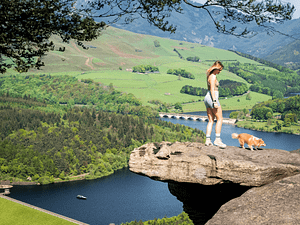 A person stood on an overhanging rock next to a small dog overlooking the British countryside.