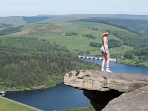 A person stood on an overhanging rock, with a lake and a bridge in the background.